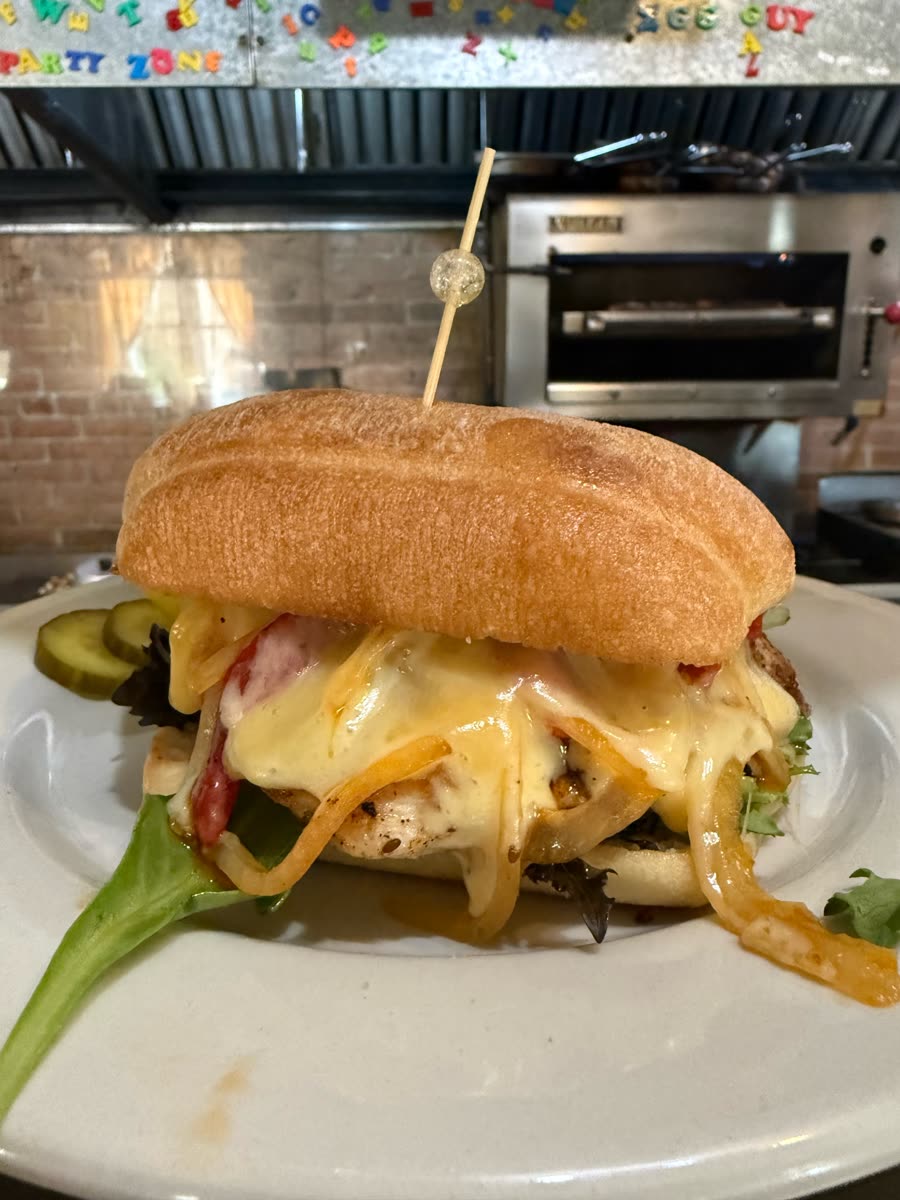 Close-up of a grilled chicken sammy loaded with melted cheese, grilled onions and tomato, with pickles on the side