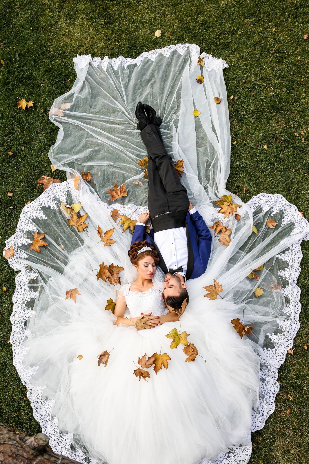 Bride and groom lying on the grass among autumn leaves, her white gown spread around them